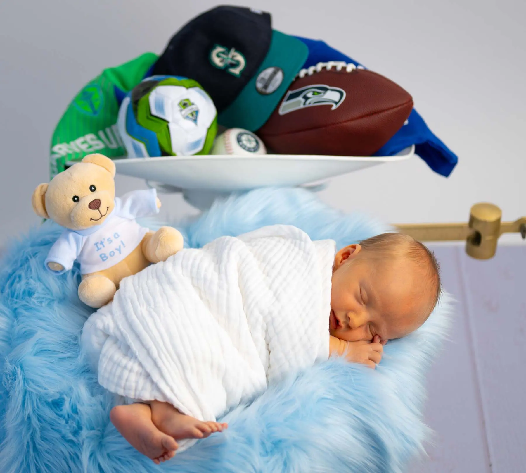 A sleeping newborn boy wrapped in a white muslin blanket lies on soft blue fur beside a teddy bear and a bowl of sports gear, conveying warmth, family pride and gentle new-parent joy.