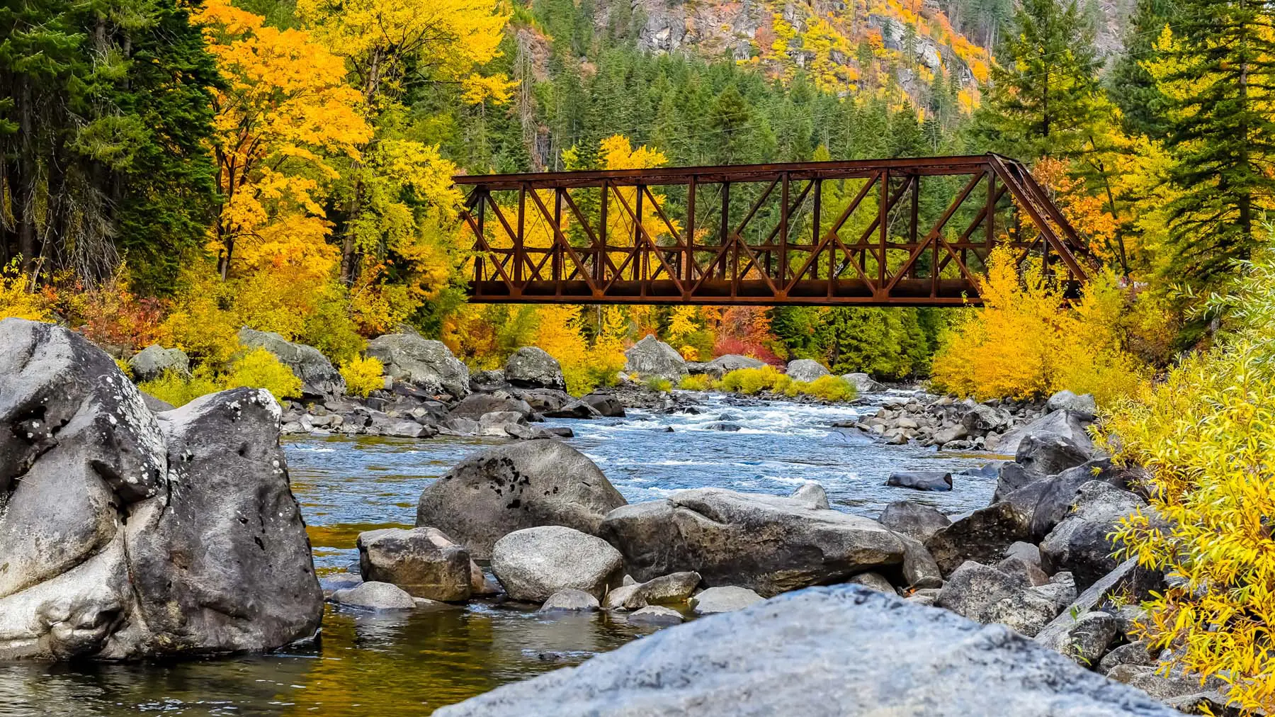 A rusty iron bridge crosses a clear mountain river framed by vibrant autumn foliage, large granite boulders and dense evergreens, creating a tranquil, scenic landscape that evokes seasonal beauty and calm.
