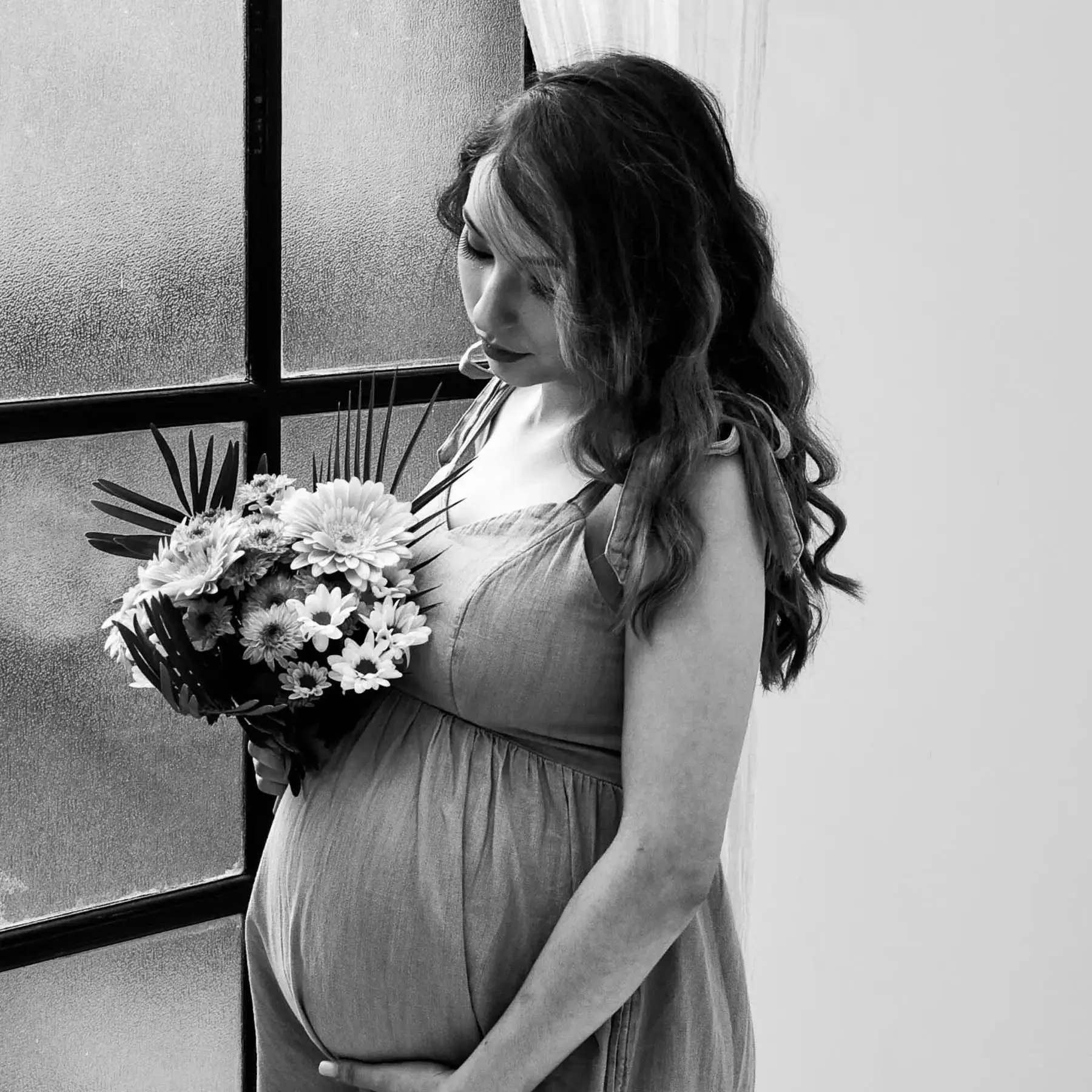 A serene maternity portrait of a pregnant woman holding a bouquet of flowers while standing by a textured window. Black-and-white tones emphasize anticipation, tenderness and the quiet connection of impending motherhood.