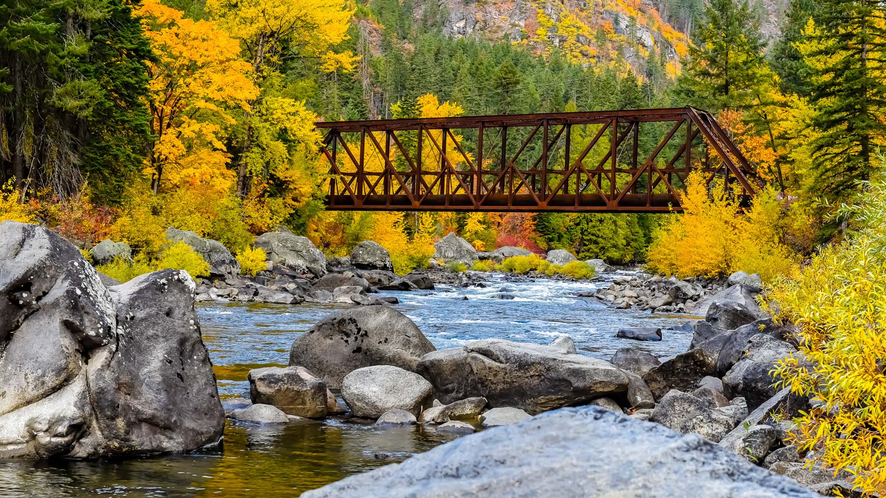 A rusty iron bridge crosses a clear mountain river framed by vibrant autumn foliage, large granite boulders and dense evergreens, creating a tranquil, scenic landscape that evokes seasonal beauty and calm.